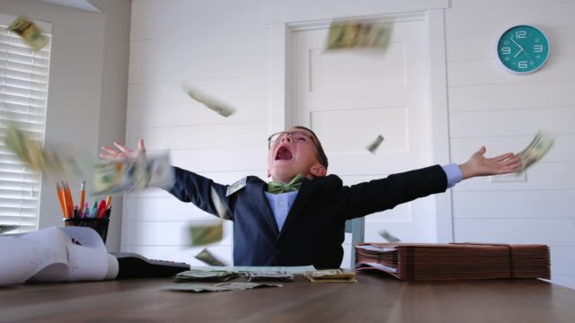 A little white kid wearing a blue suite, sitting at a desk with a calculator, pen and pencils inside of holder and a folder on top of the desk throwing money in the air.