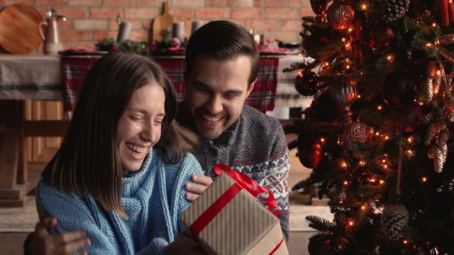 a white couple opening a Christmas gift together in front of the Christmas tree