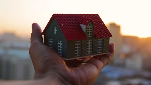 A white person holding a miniature green house with a red roof and white pained windows. 