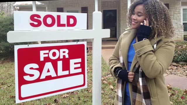 A black girl with a camel coat with a black, white, brown and grey scarf, on the phone standing in front of a brick house with a sold/ for sale sign in the front lawn.