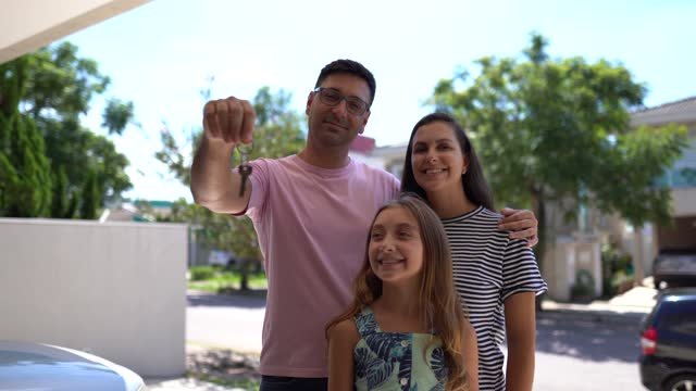 A family (Father, mother and daughter) standing in front of their new home.  The father is holding the front door keys in his hands. 