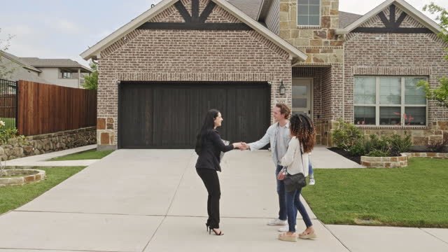 A realtor shaking hands with a couple in front of a beige brick home.  They are shaking hands.