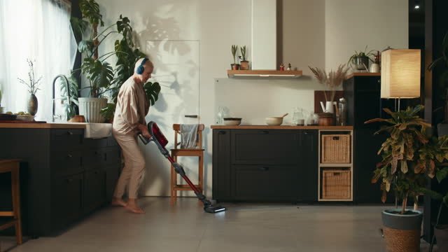 A male in a beige outfit, wearing headphones, vacuuming the kitchen floor with a bunch of flowers 