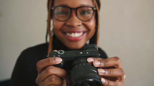 A black lady holding a camera wearing black round glasses and a black sweater. 