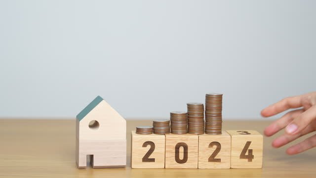 A white hand reaching for 2024 wooden building blocks with coins on top of them next to a wooden mini house.