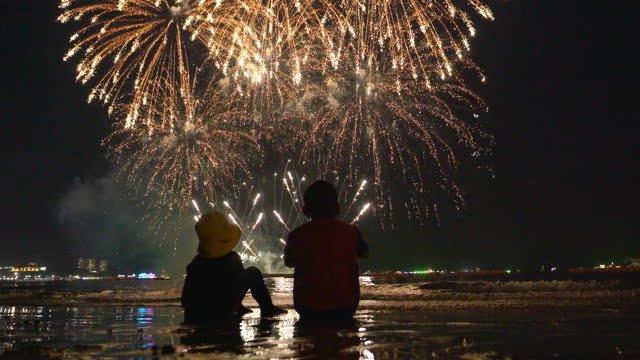 A couple sitting next to a body of water looking at fireworks. 