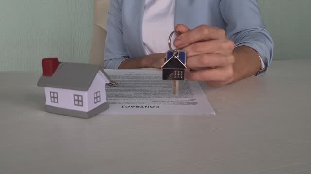 A white male holding house keys, sitting at a table with a contract and a miniature house sitting on it.