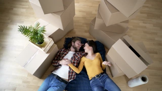 A white couple daydreaming about their future laying on the floor in a large room with boxes  and a potted palm tree.stacked around them